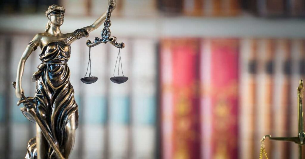 A brass statue of Lady Justice with her scale with lawbooks behind her